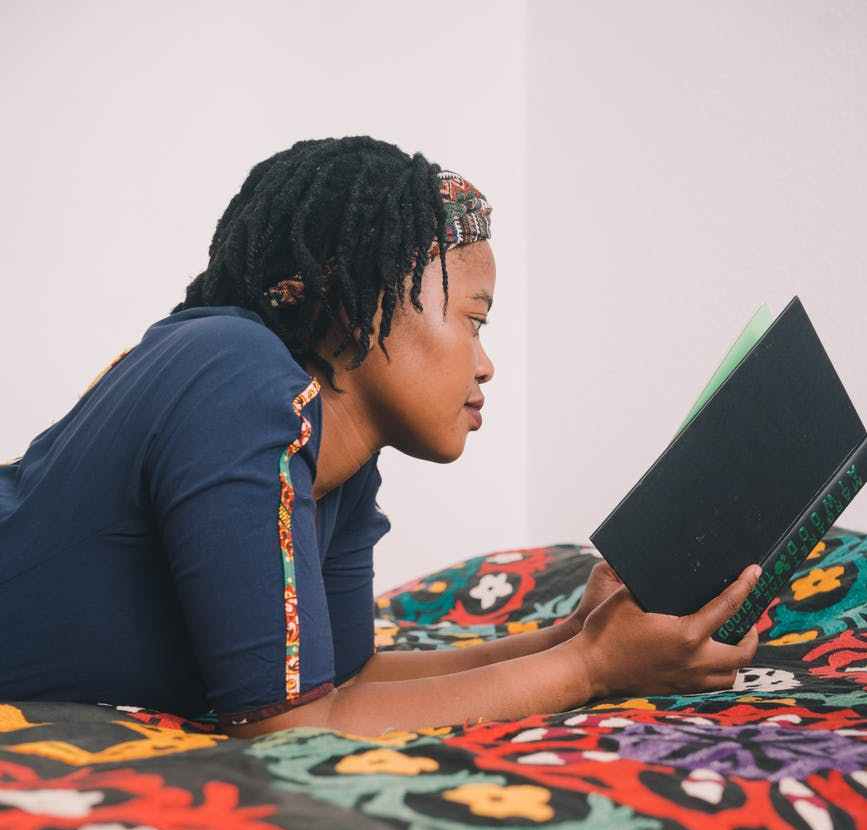 Black woman reading green hardcover book while lying on colorful bedspread