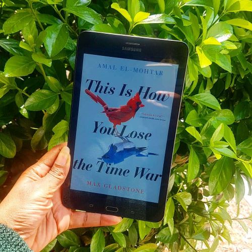 ID: A black tablet displaying the cover of This Is How You Lose The Time War(sky blue background with red and blue cardinal birds reflecting each other surrounded by title text). The tablet is held in front of a leafy green bush by a black woman's hand.