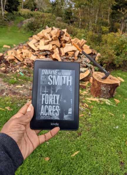 A Black woman's hand(my hand) holds a Kindle displaying the black and white cover of Forty Acres in front of a freshly chopped woodpile, complete with axe.
