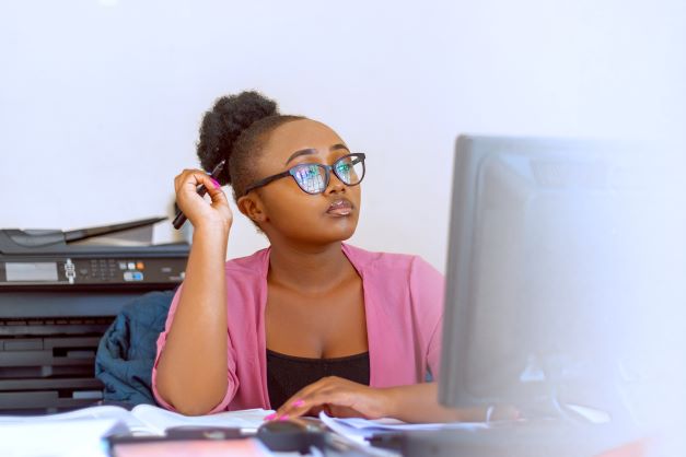A cute Black woman with glasses, an Afro-puff, wearing a pink cardigan and a black shirt, is sitting in front of a computer monitor deep in thought.