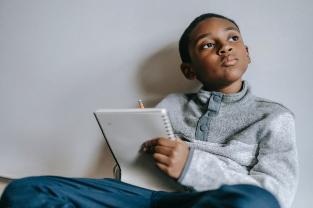A young Black boy sits against a white wall with a notebook and pencil in his hands, staring into space pensively