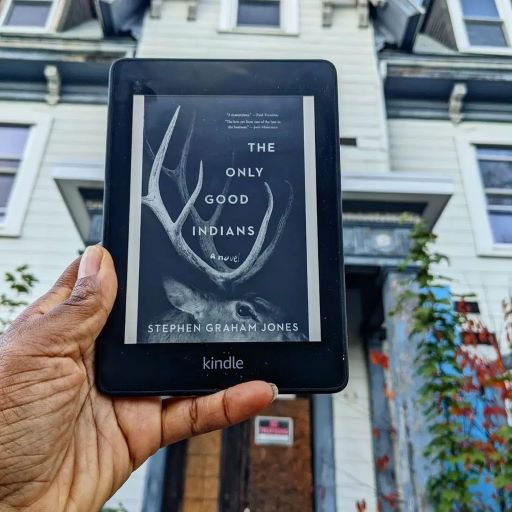 My hand holds up a Kindle displaying the cover of The Only Good Indians--a mysterious pair of antlers in the dark. In the background, there is a huge and creepy old house, a condemned sign barely visible.