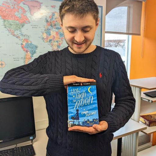 A young Ukrainian man in a Black sweater smiles down at the blue and gold cover of How High The Moon. Behind him are a world map, an orange map, and an open window looking out onto a public square.