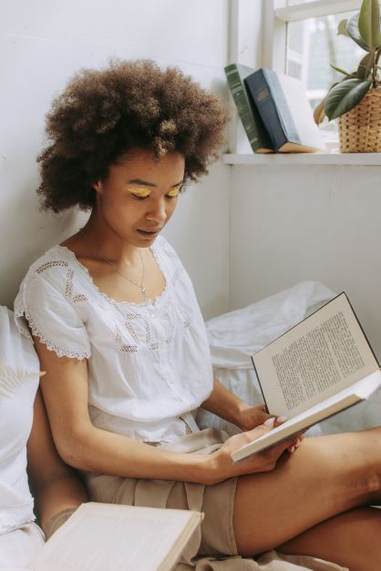 A young Black woman with a big afro and bright yellow eyeshadow sits in a cozy corner,reading a hardcover book.