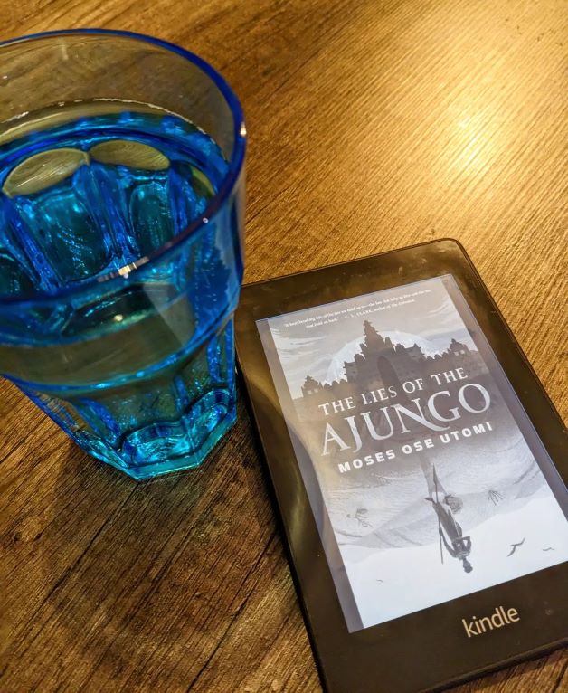A Kindle displaying the cover of The Lies of the Ajungo lies on a kitchen table next to a full blue glass of water.