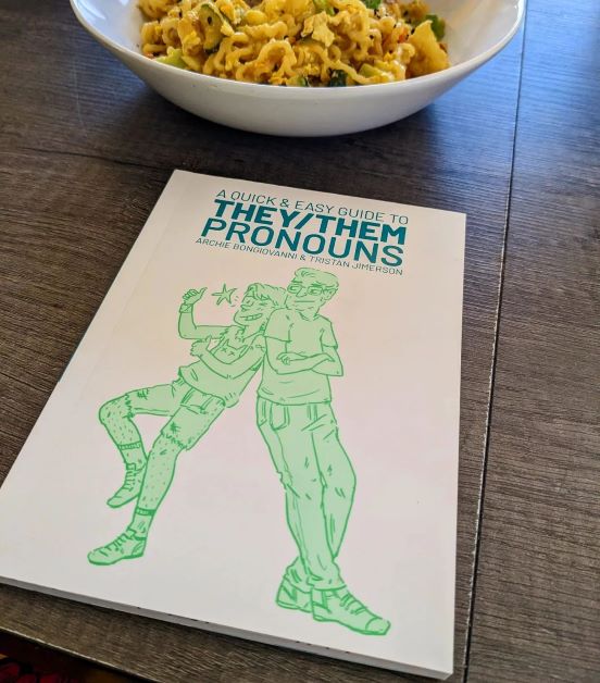A Quick and Easy Guide To They/Them Pronouns lies on a wooden kitchen table in front of a white bowl heaped with delicious egg noodles and veg.