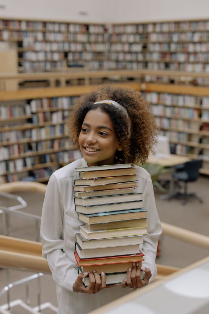 A cute black girl with a fluffy afro holds a tall stack of books in a library.
