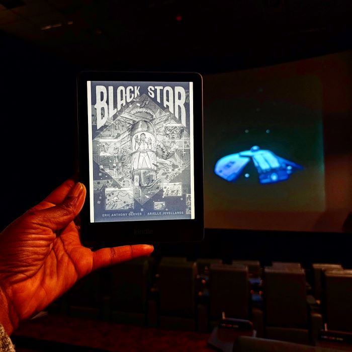 The Kindle cover of Black Star, depicting a woman trapped in a glowing space tube, is held up in a dark movie theater in front of a projection screen. The movie playing is the classic camp film Dark Star.