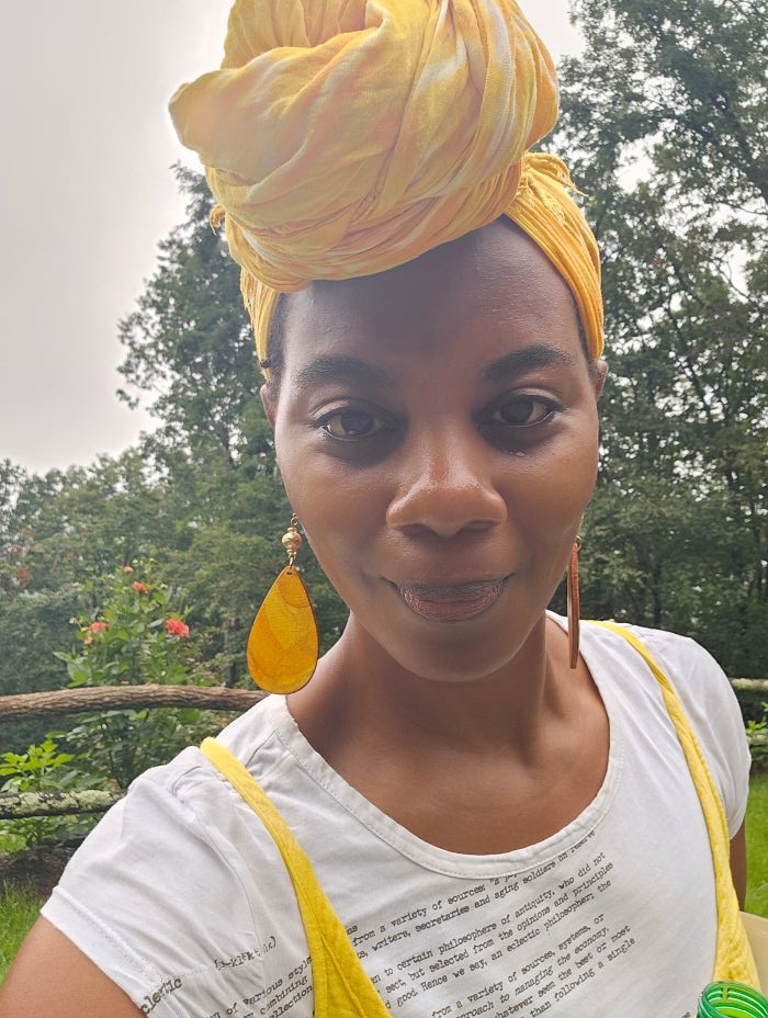 The writer, a Black woman in a tall yellow headwrap, a yellow spaghetti strap dress and a t-shirt with the dictionary definition of "eclectic" printed on it, gazes playfully down at the camera.