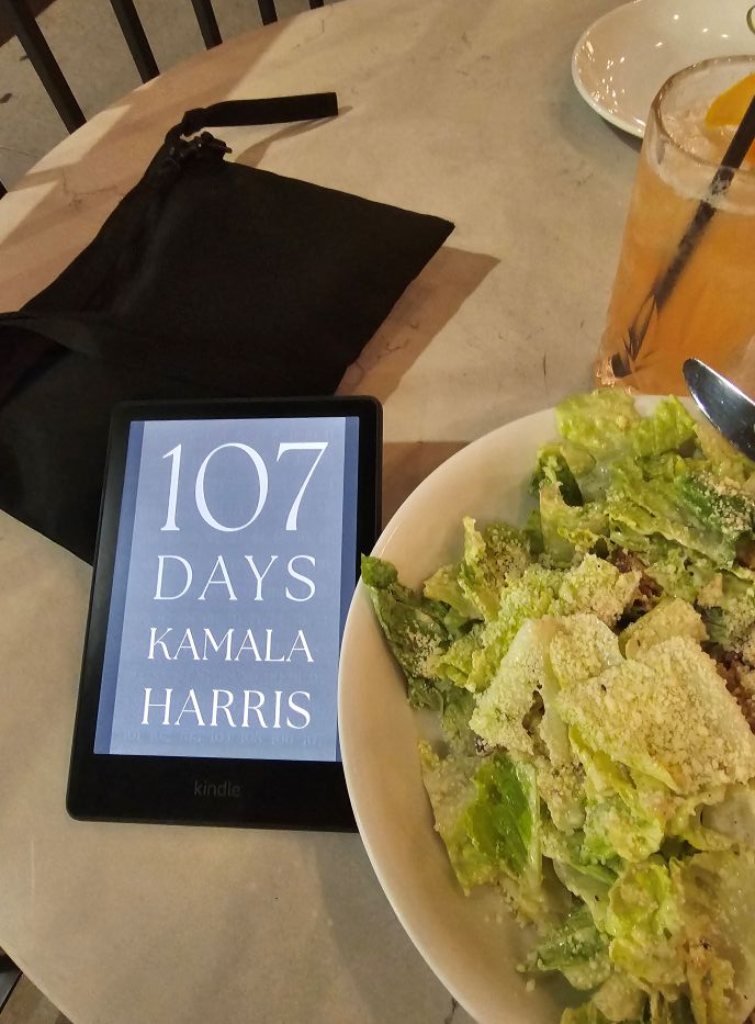 The Kindle cover of Kamala Harris' memoir, displaying only the words "107 Days" in plain text, sits next to a wide bowl of Caesar salad and a glass of beer.