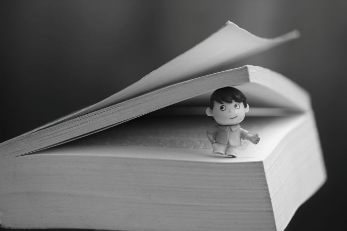 Black and white photograph of a tiny light skinned boy with short dark hair standing between the open pages of a thick book.