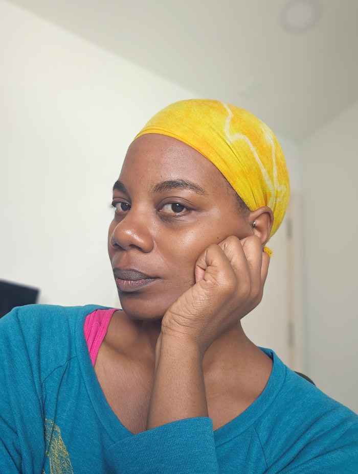 The author, a tired Black woman, is in three-quarter profile to the camera, seen from the shoulders up, wearing a slouchy turquoise sweatshirt and a yellow headwrap