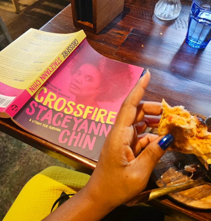 The paperback cover of Crossfire, displaying young StacyAnn Chin and her glorious fro in profile on the front, is face down on a wooden table. Next to it is a Black woman's hand(my hand) holding a half-eaten empanada.