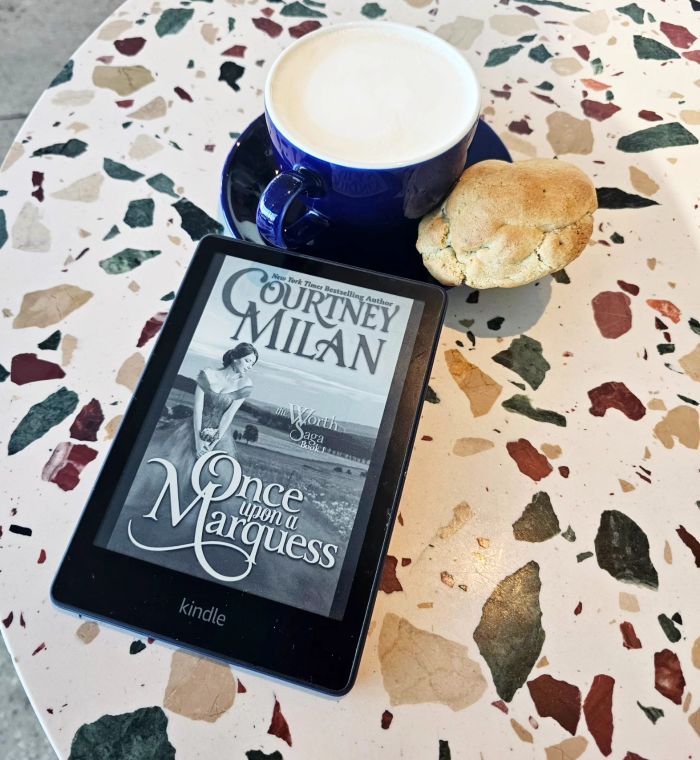 The Kindle cover of Once Upon A Marquess depicts a young white woman in a Victorian dress sitting in a grassy field. The Kindle lies on a speckled ceramic table, nest to a saucer with a blue ceramic cup of milky coffee and a half-eaten scone on it.