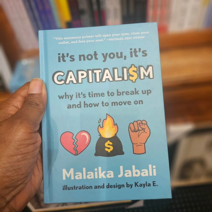 The blue hardback cover of It's Not You, It's Capitalism is held up by a Black woman's hand in front of a loaded bookshelf. The cover depicts a series of illustrated emoticons: a broken heart, a bag of money on fire, and an upraised, clenched Black Power fist.