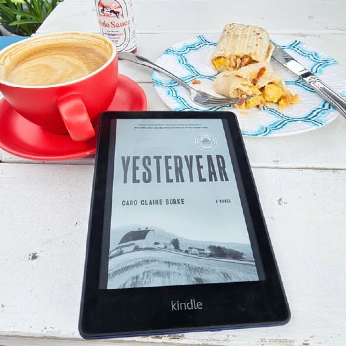 The black and white Kindle cover of Yesteryear, depicting a bleak frontier landscape, lies on a white wooden picnic table near a red ceramic cup of coffee and a half-eaten breakfast burrito.