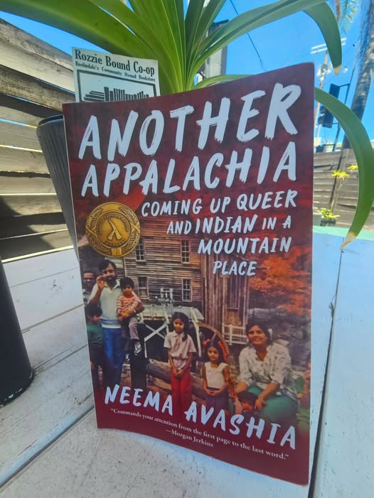 The paperback of Anofher Appalachia rests against a potted plant. The cover depicts an Indian family sitting in front of a clapboard house.
