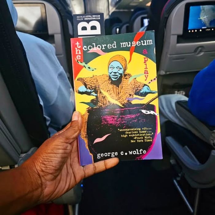 The colorful cover of the play The Colored Museum, depicting a Black woman in a headscarf stirring a cauldron, is held u in the aisle of a busy commercial flight. Sorry to my fellow passengers.