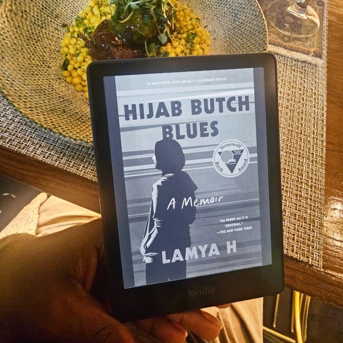 Mel's hand props up a Kindle displaying the black and white cover of Hijab Butch Blues, which depicts a slim hijabi in baggy trousers and shirt facing away over an abstract line art landscape. The Kindle rests against a delicious dish of couscous and braised beef in a wide painted ceramic dish.