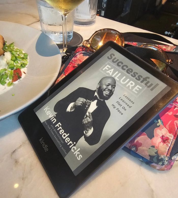 The Kindle cover of Successful Failure, depicting comedian Kevin Fredericks power posing in a tux, lays against a flowered wallet on a dining table. Behind it are a dish of salad and a glass of white wine.